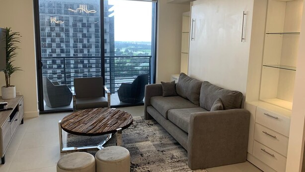 Una sala de estar moderna con un sofá, un sillón y otomanas, una mesa de centro de vidrio y vistas a un paisaje urbano a través de ventanas amplias.