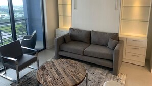 Un salón moderno con un sofá gris, una mesa redonda de centro y vistas a un paisaje urbano a través de grandes ventanales.