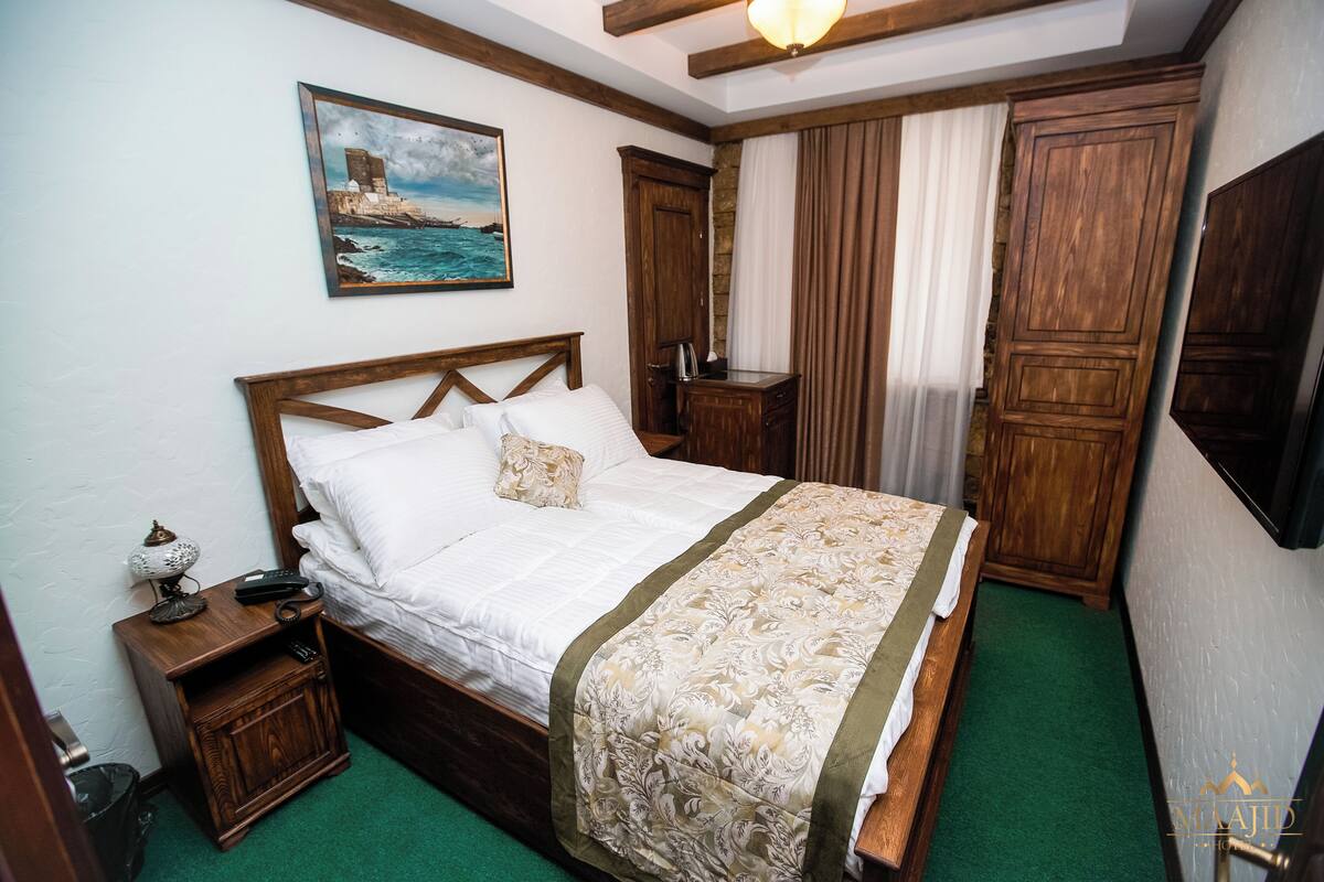 Una cama bien hecha con una colcha estampada, una mesita de noche con una lámpara, una mesita auxiliar de madera y un cuadro de una escena costera en la pared.