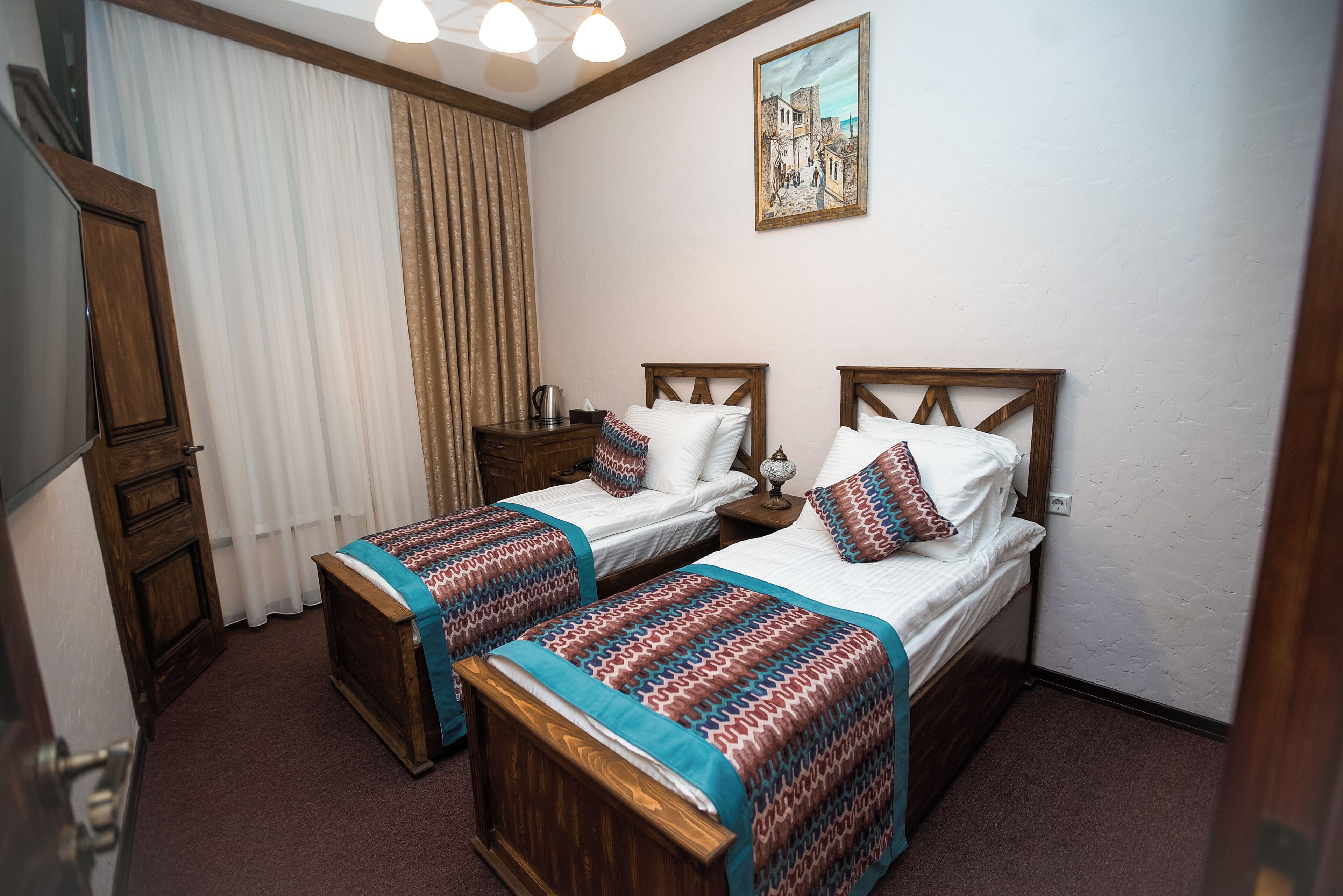 A hotel room with two wooden beds, a wooden wardrobe, a painting on the wall, and a window with curtains.