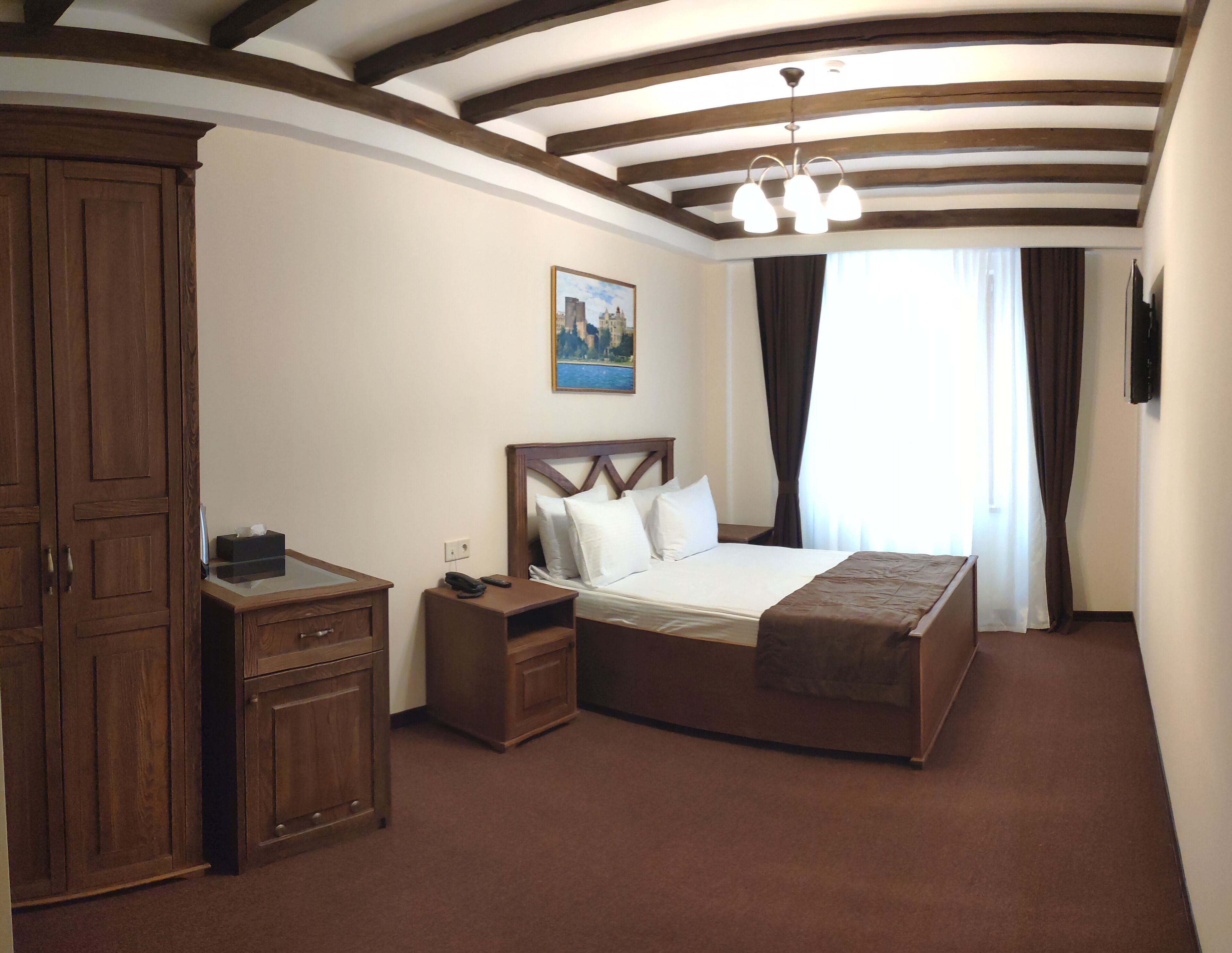 A hotel room with a large bed, wooden wardrobe, bedside table, and a painting on the wall.