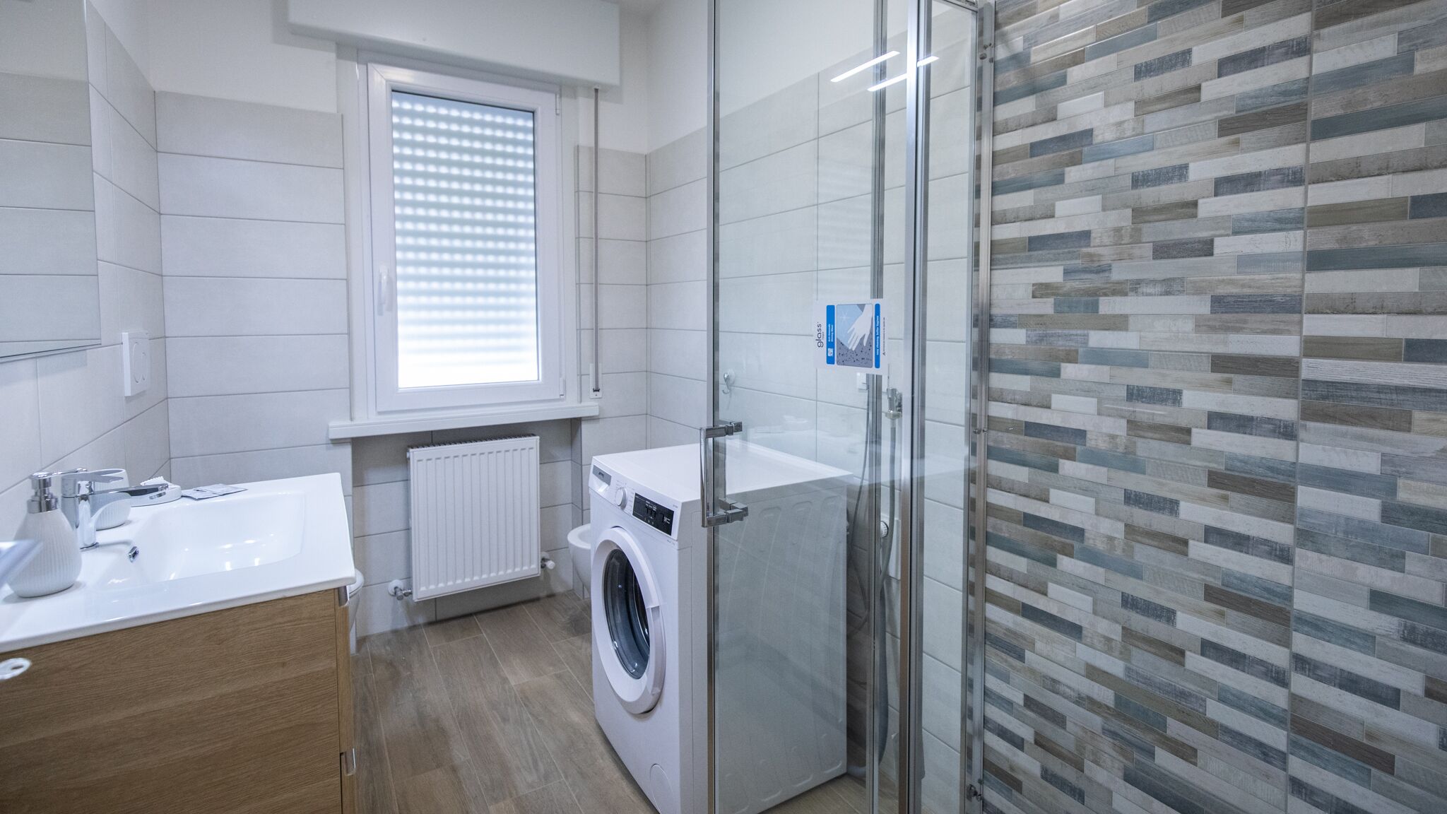 A modern bathroom with a washing machine, a glass shower enclosure, and a window with blinds.