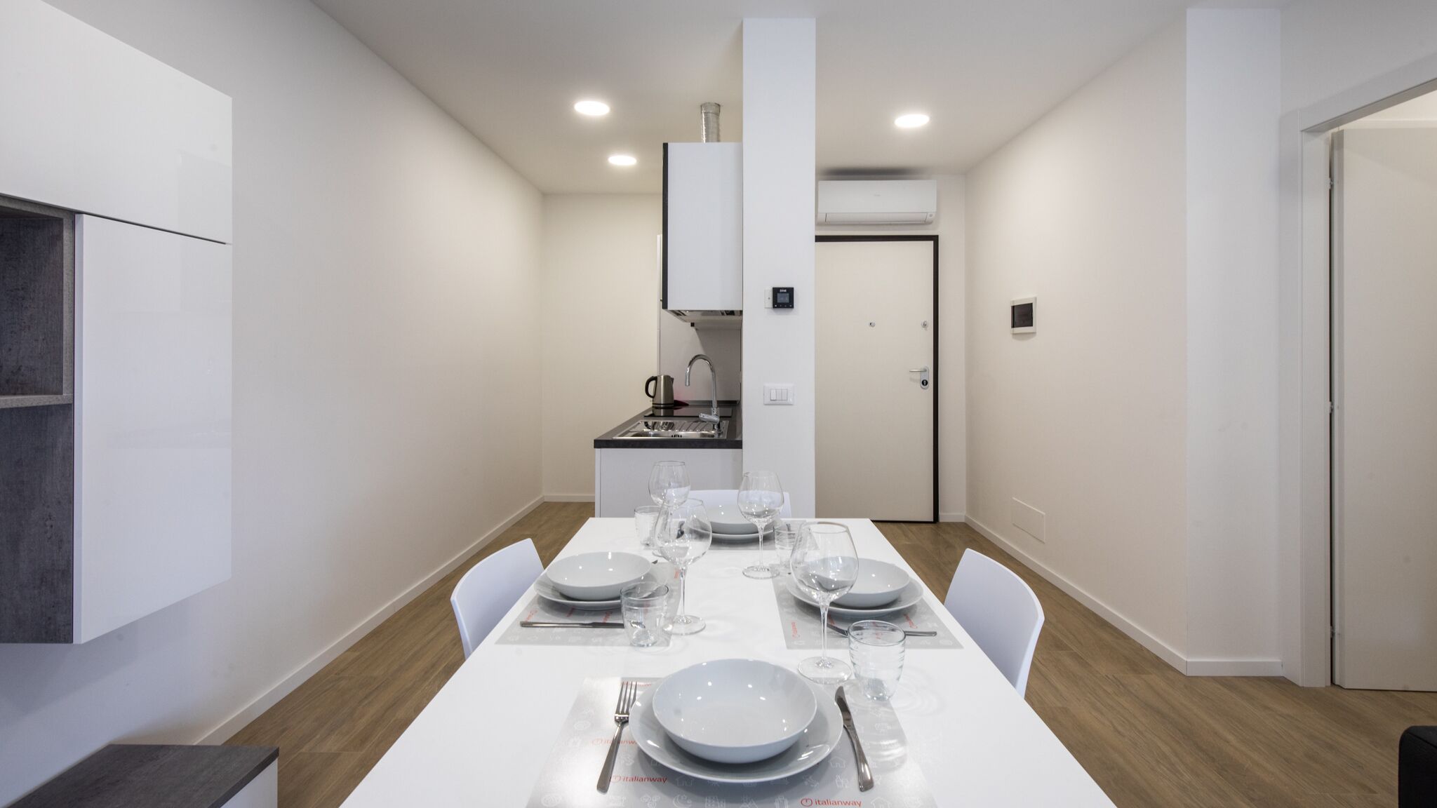 Un espace repas moderne avec une table blanche dressée pour quatre personnes, une cuisine en arrière-plan et une porte donnant sur une autre pièce.