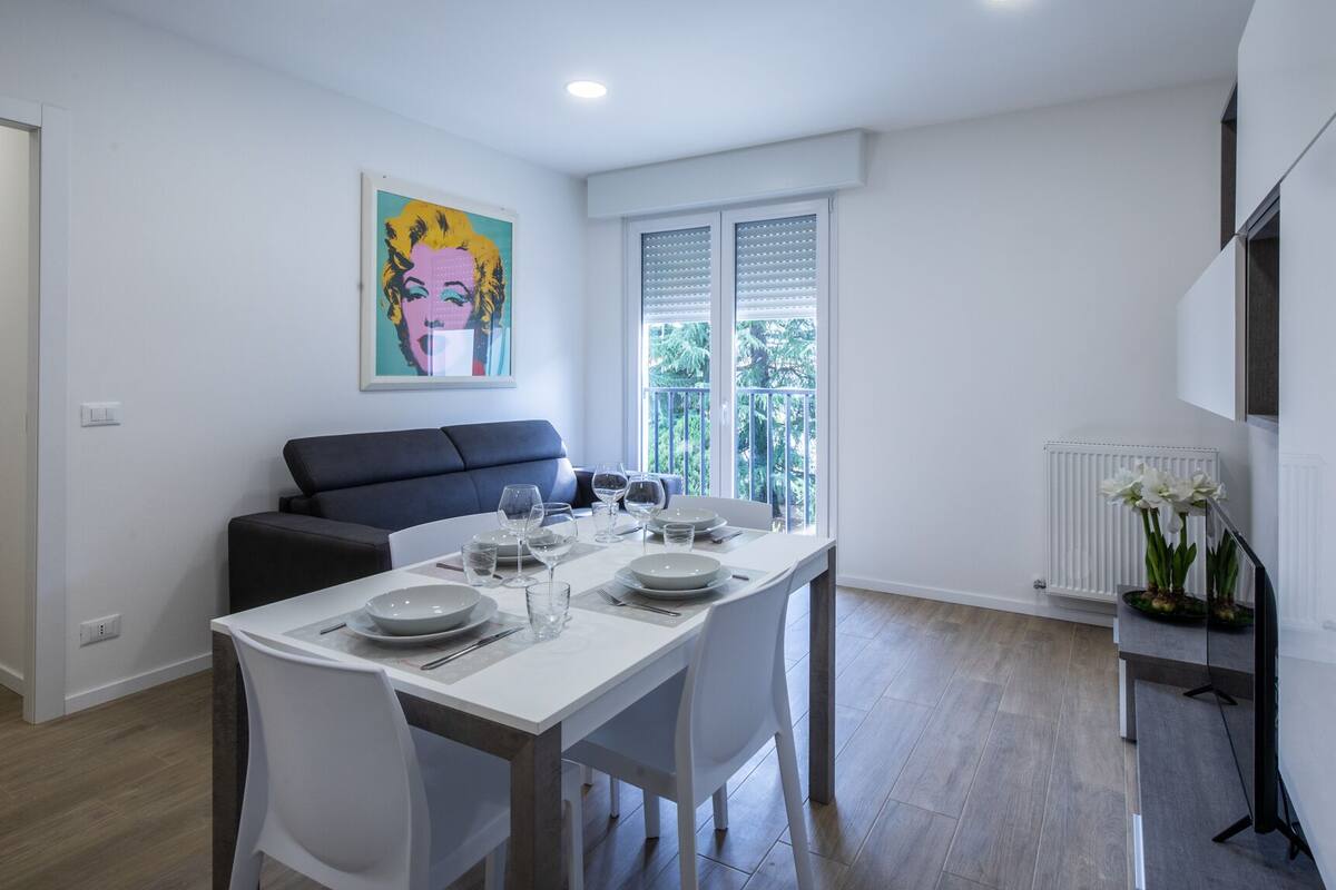 A dining area with a white table set for four, a grey sofa, and a large window with blinds.