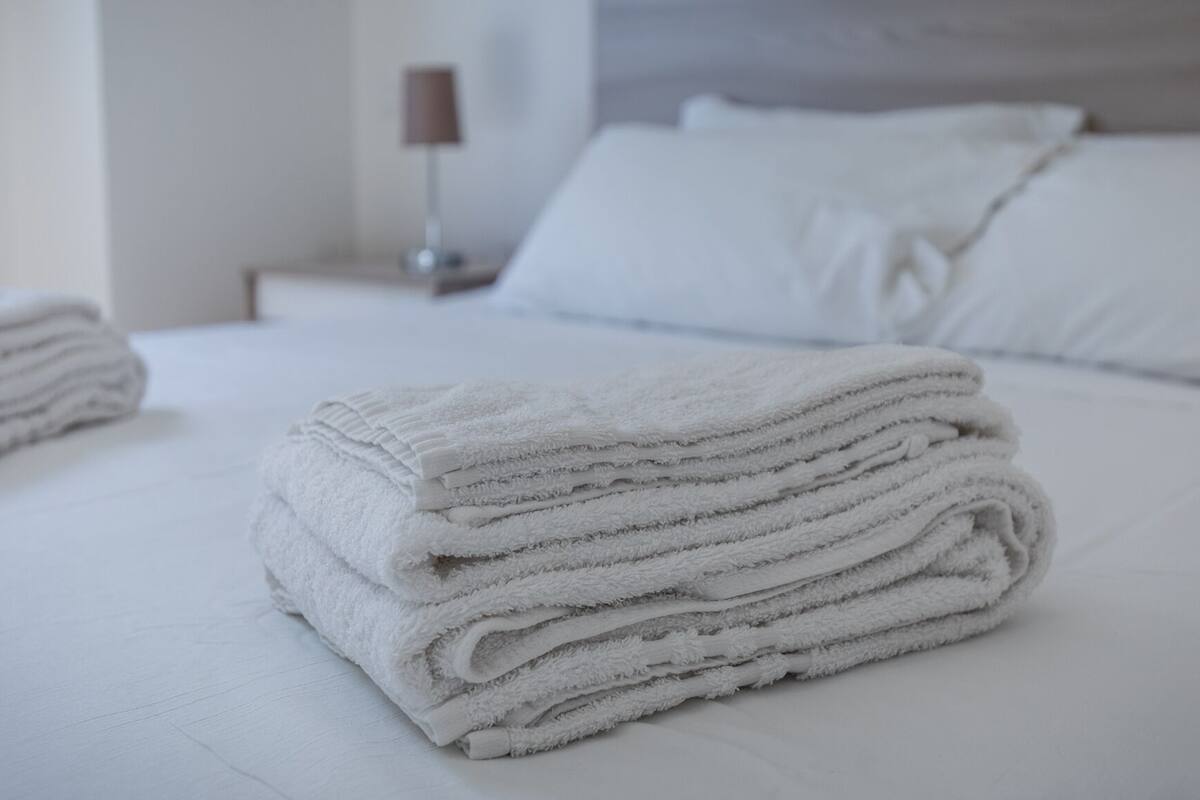 A neatly made bed with white linens and pillows, a bedside table with a lamp, and a wall-mounted shelf.