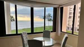 Una habitación con una mesa redonda de vidrio y cuatro sillas, ventanas amplias con vistas al mar y palmeras, y un balcón con vista a un edificio.