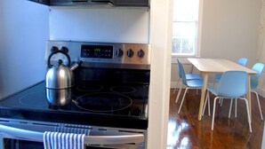 Une cuisine compacte équipée d’une cuisinière, d’un four et d’une bouilloire. On y trouve une table à manger avec des chaises, ainsi qu’une fenêtre avec des stores.