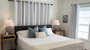 Un dormitorio con una cama grande, dos mesitas de noche con lámparas, un ventilador de techo y una ventana con cortinas.