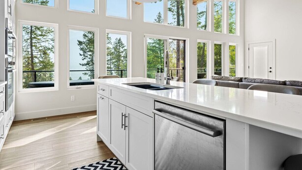 A modern kitchen with a large island, white cabinetry, and multiple windows offering views of trees.
