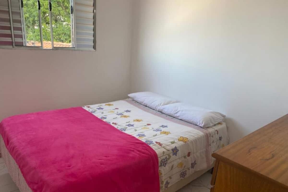 A single bed with a floral bedspread and a pink blanket, a wooden nightstand, and a window with blinds.