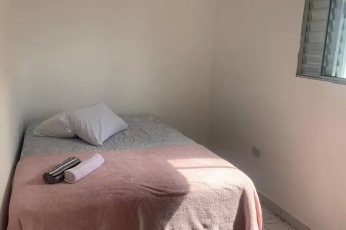 A small, neatly made bed with a pink blanket and a rolled towel, a white pillow, and a window with blinds.