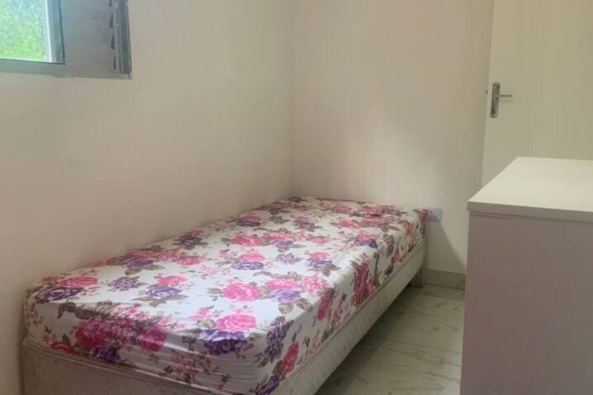 A small, narrow room with a single bed covered in a floral bedspread, a white cabinet, and a door.