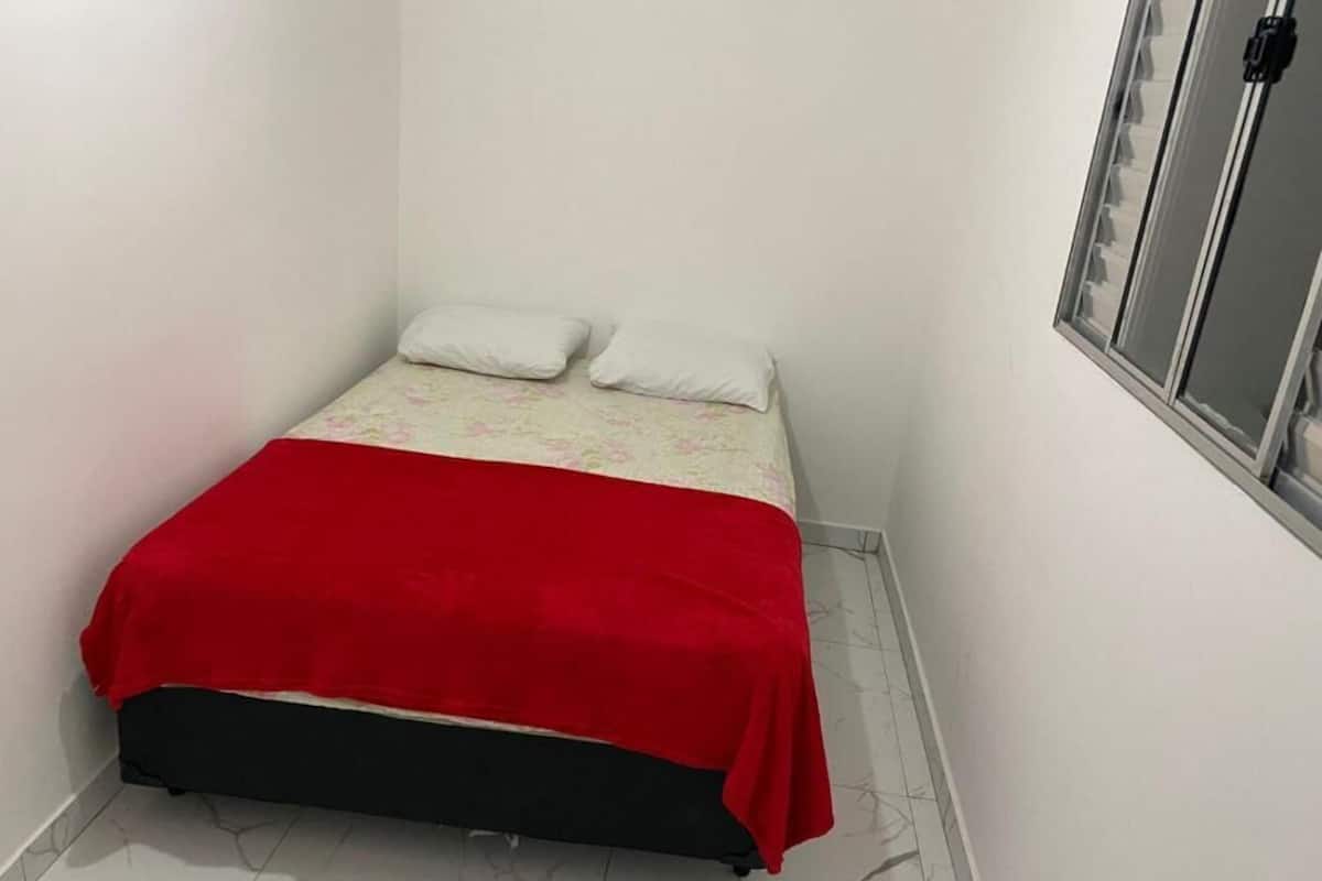A small, single-bed room with a red bedspread, white pillows, and a window with a metal frame.