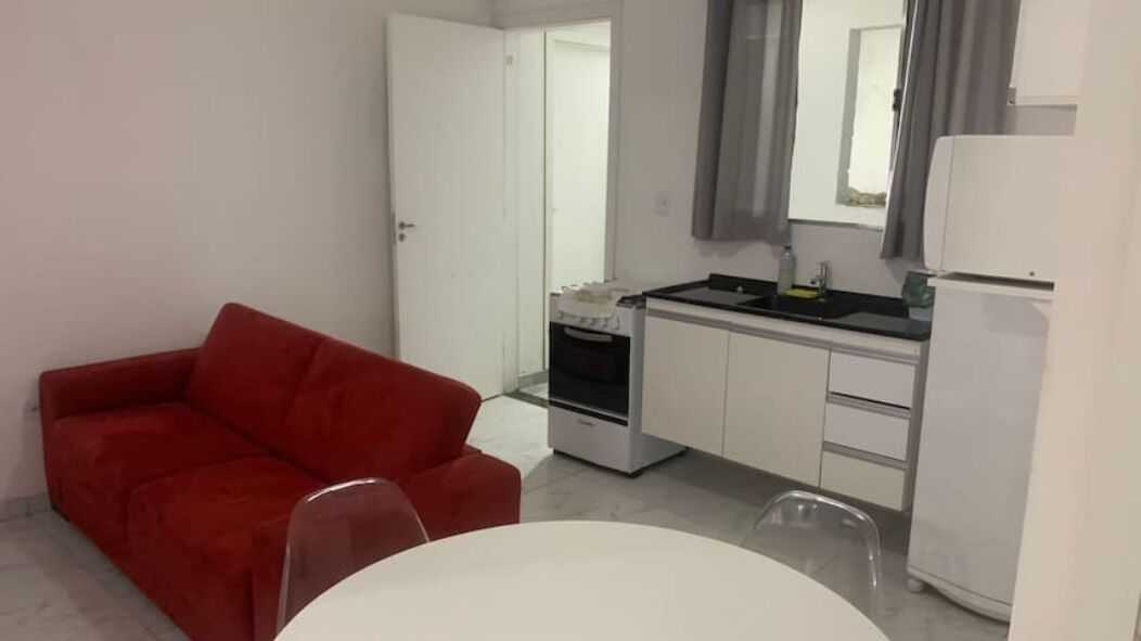 Een ruimtelijke leefruimte met een rode bank, een witte eettafel, een kitchenette met fornuis en een koelkast.