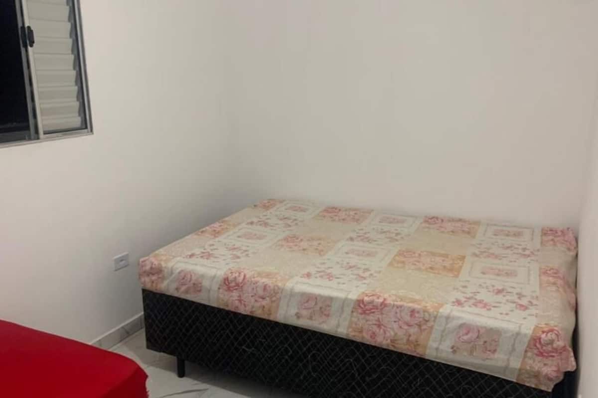A small, single-bed room with a floral bedspread, a red bench, and a window with blinds.