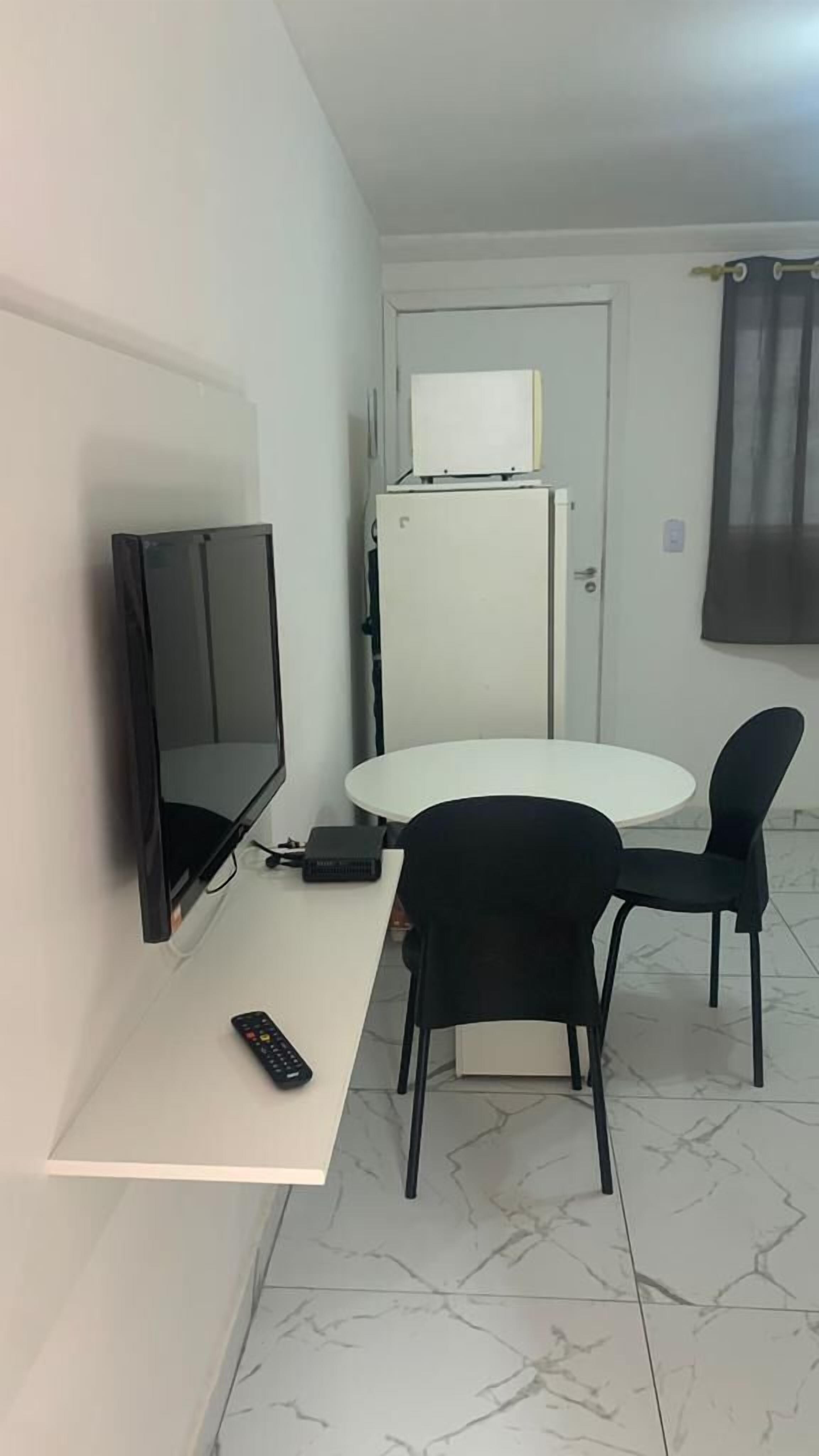 Ein kleines Zimmer mit weißer Wand, einem an der Wand befestigten Flachbildfernseher, einem weißen Tisch mit zwei schwarzen Stühlen, einem weißen Kühlschrank und einer Tür mit Vorhang.