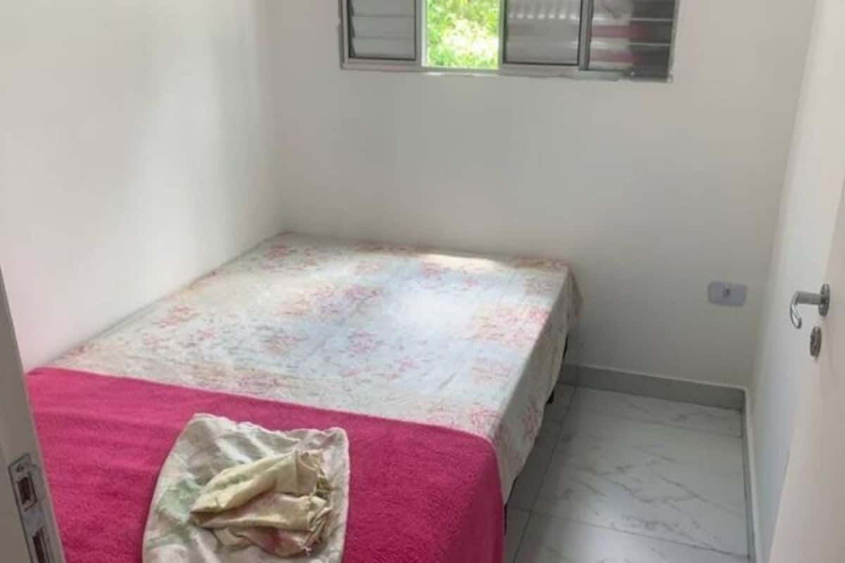 A small, single-bed room with a floral bedspread, a pink towel, and a window with blinds.