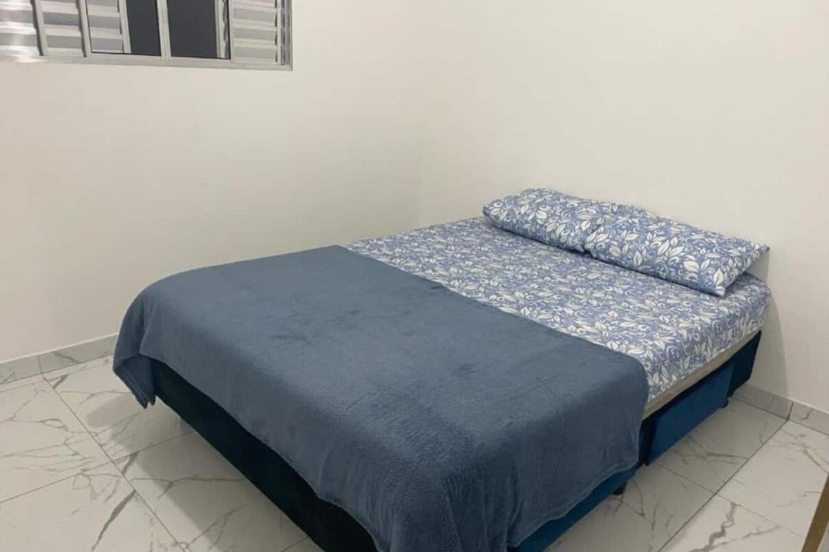 A single bed with a blue bedspread and matching pillow, a blue blanket, and a window with blinds.