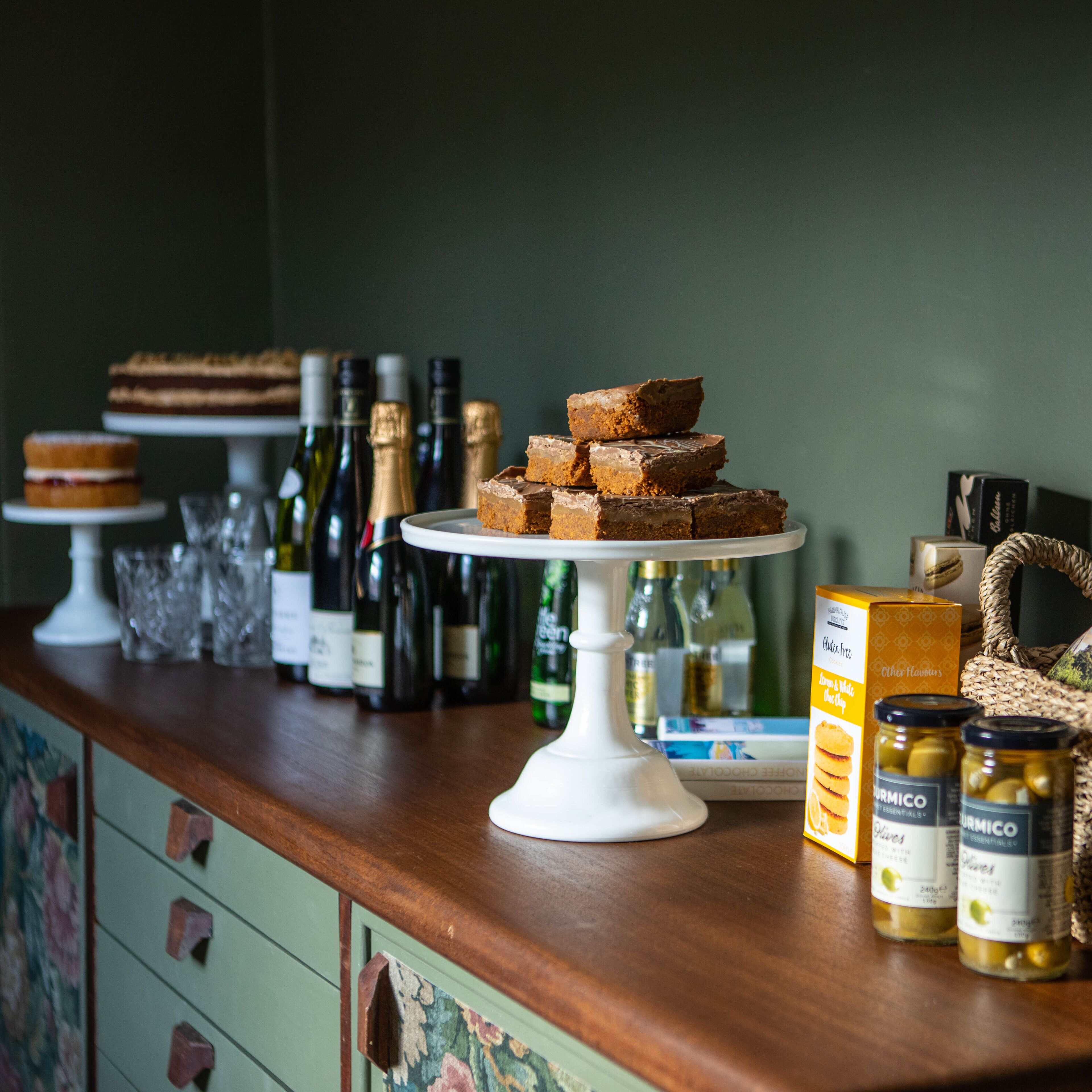 A dessert display with various cakes and pastries on a wooden counter, surrounded by wine bottles and jars of preserves.