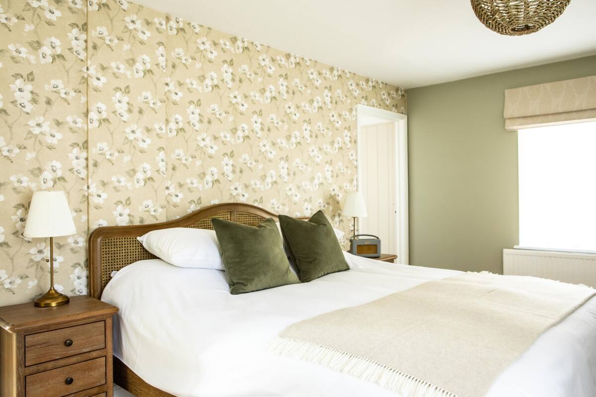Una cama bien tendida con sábanas blancas y dos almohadas verdes, una lámpara de noche y un estampado de flores en el papel tapiz.