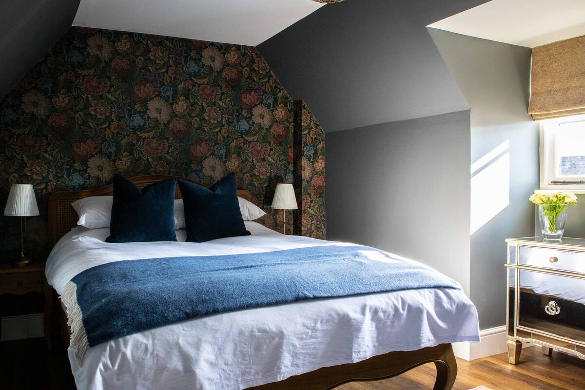 Un dormitorio con una cama grande, dos mesitas de noche, una mesita auxiliar, un papel tapiz con estampado floral y una ventana con vistas.