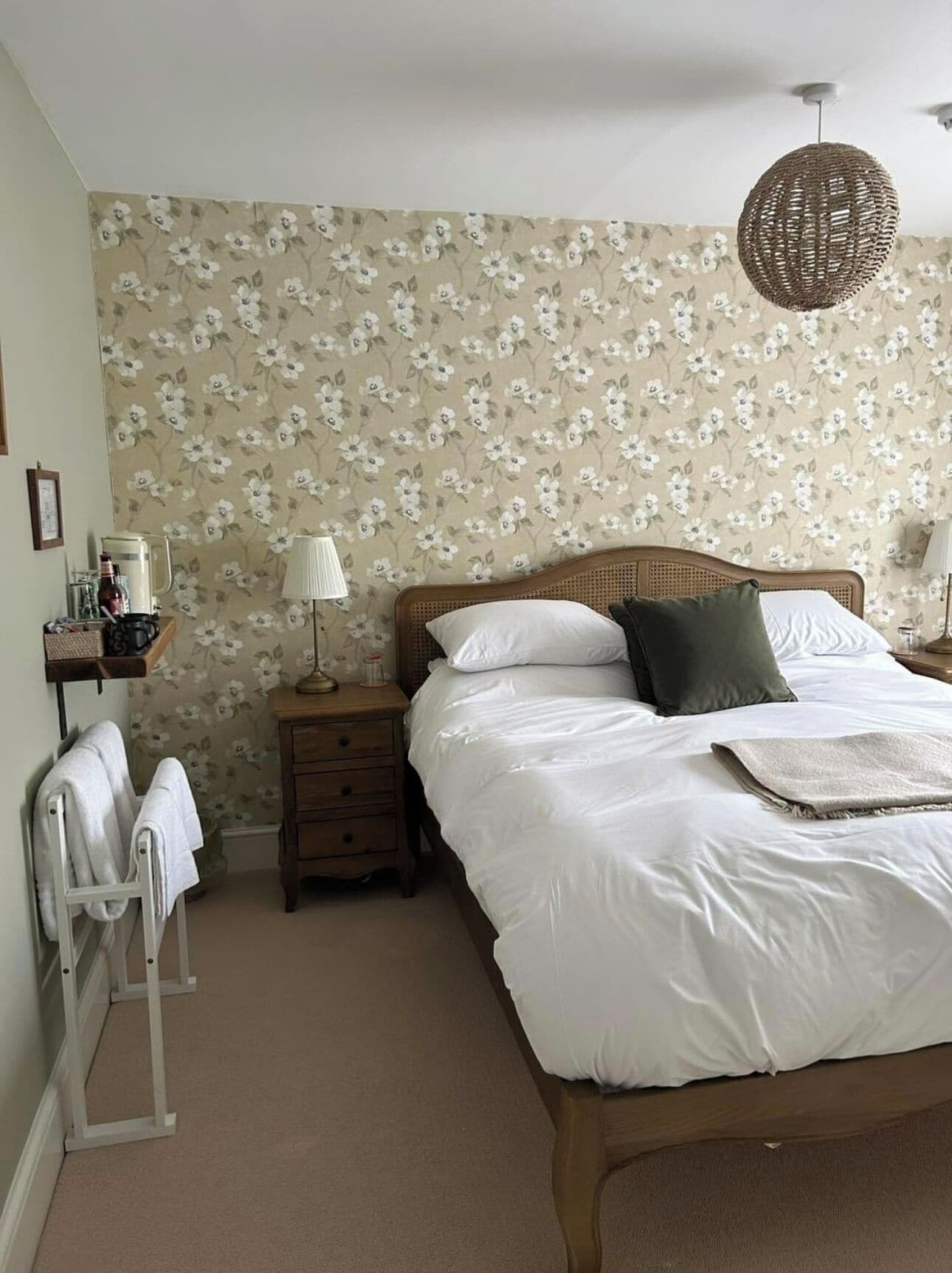 A bedroom with a bed, a nightstand, a lamp, a wall with floral wallpaper, and a hanging light fixture.