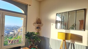 A room with a view of a cityscape through a large window, a potted plant on a wooden table, a lamp, and a wall-mounted shelf with decorative items.