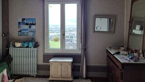 A room with a large window, a radiator, a small cabinet, a mirror, and a wall with a collage of photos.