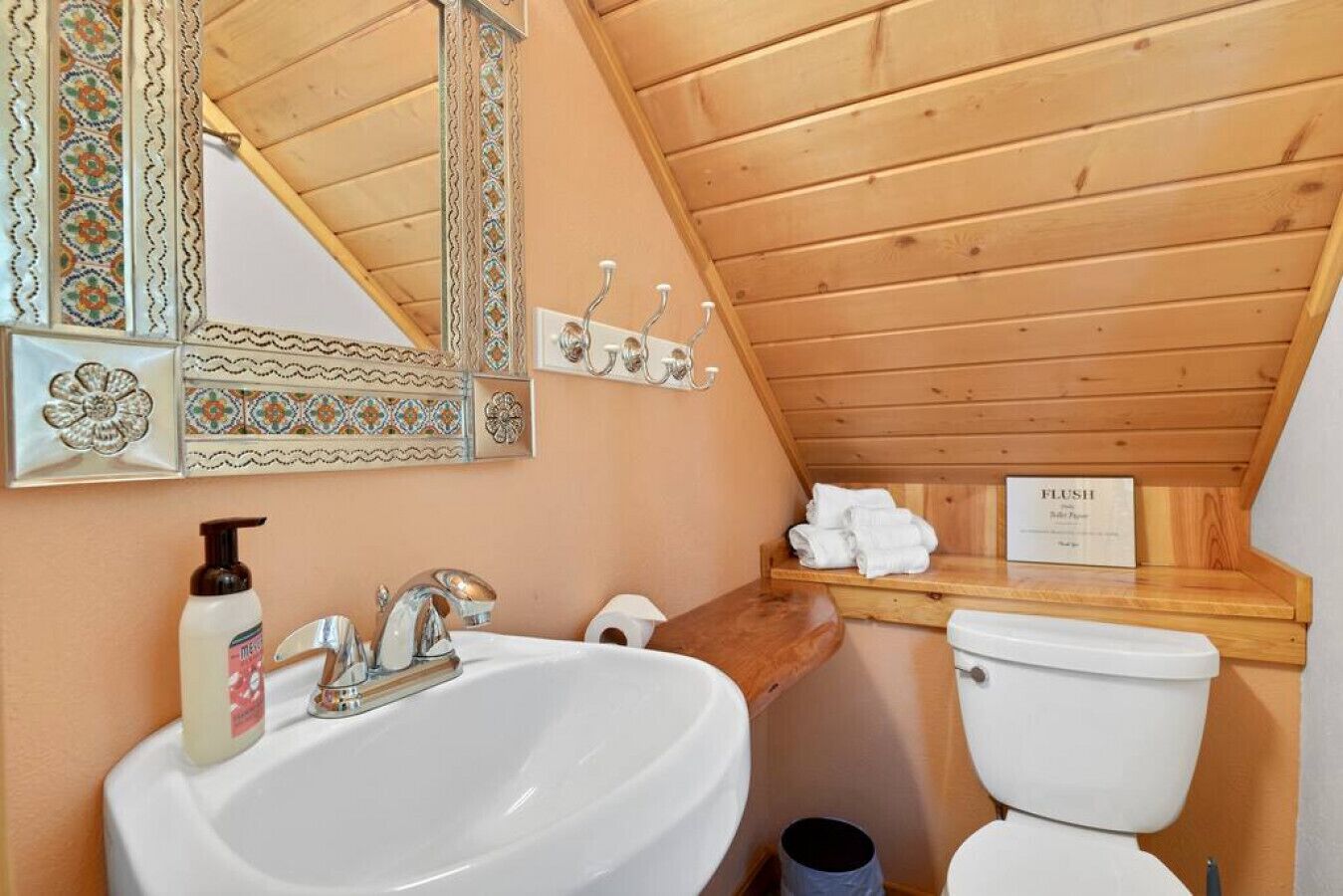 A small bathroom with a wooden ceiling, a sink, a toilet, and a mirror with decorative tiles.