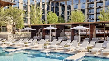 A modern hotel pool area with sun loungers and umbrellas, surrounded by a multi-story building with large windows and trees.