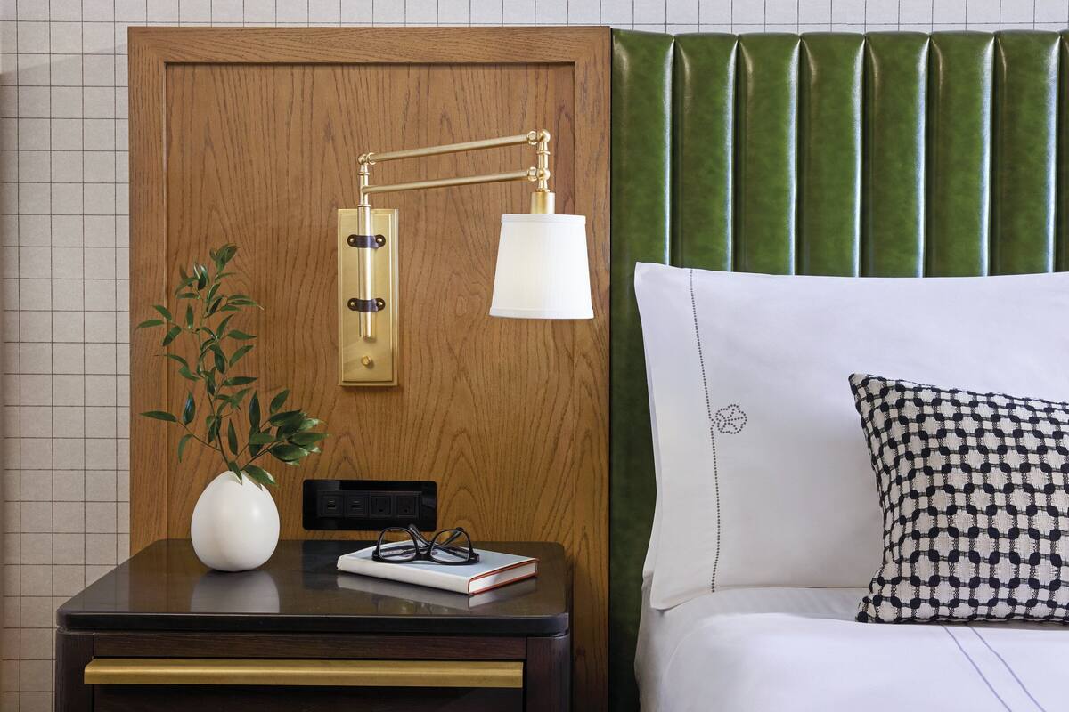 A neatly arranged hotel room with a wooden headboard, a bedside table with a lamp, a plant, and a book.
