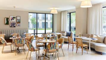 A modern hotel room with a dining area, a sofa, and large windows offering a view of the city.