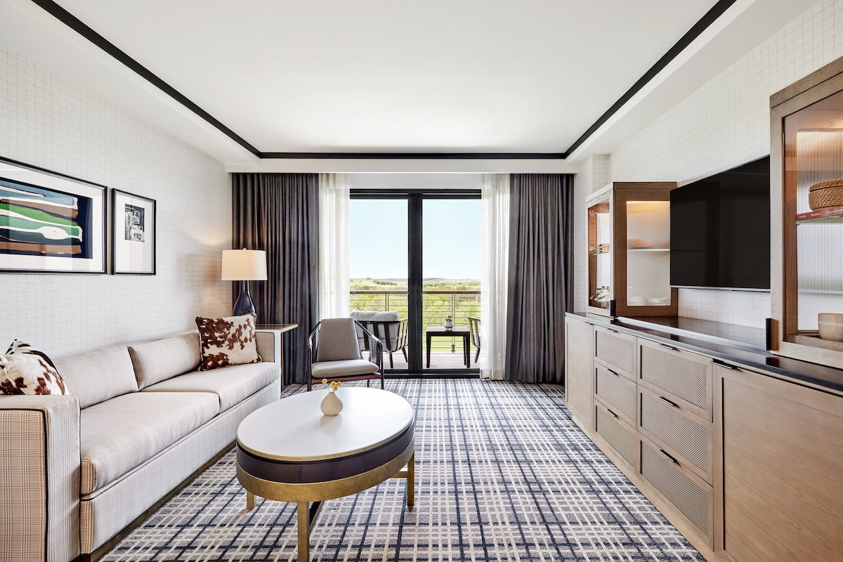 A hotel room with a sofa, a round table, a TV, a balcony with a view, and a patterned carpet.