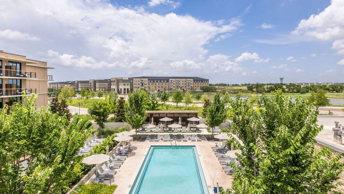 A swimming pool with lounge chairs and umbrellas, surrounded by trees and a building in the background.