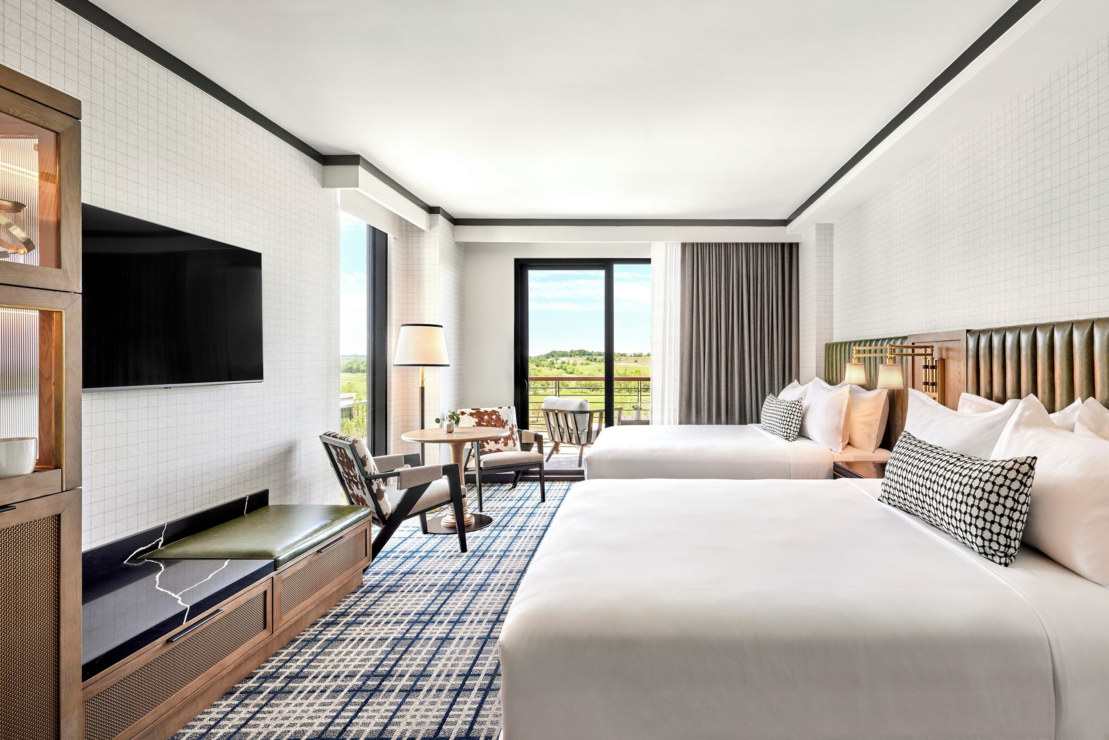A hotel room with two beds, a TV, a dining area, and a view of the outdoors.