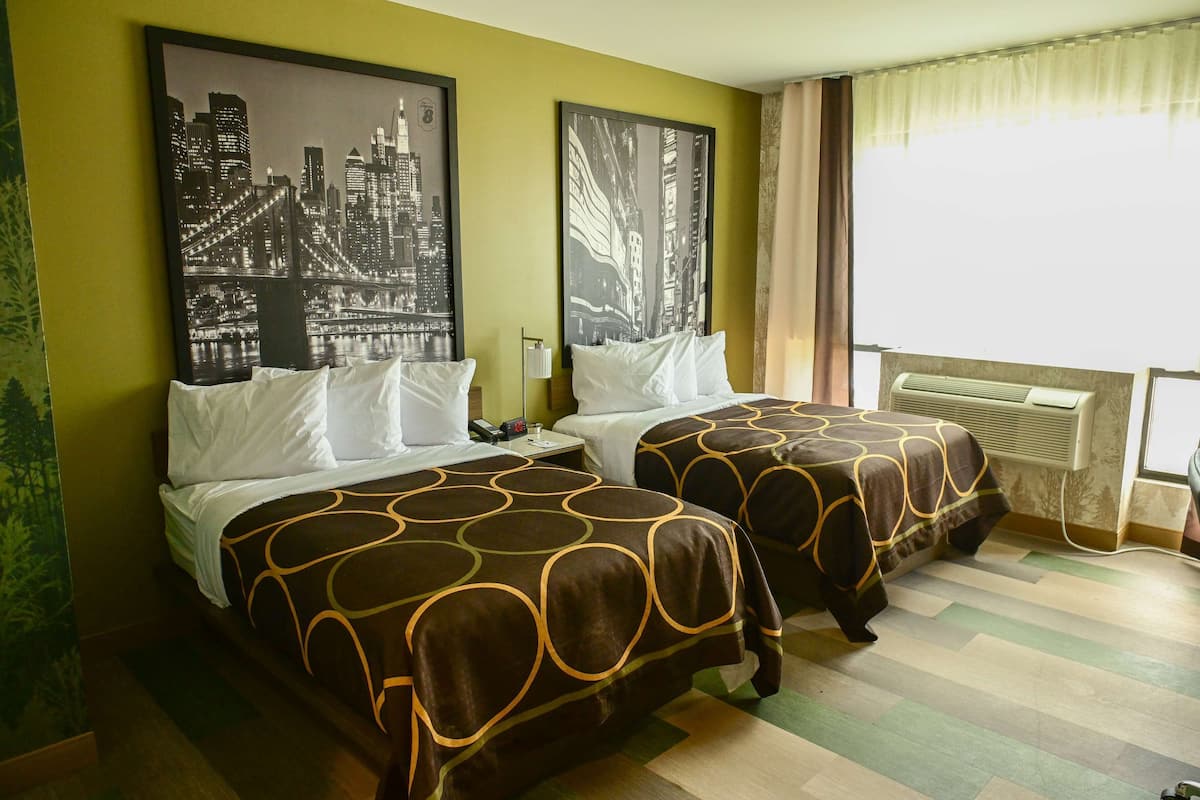 A hotel room with two beds, each with a patterned bedspread, a large window with curtains, and a wall-mounted air conditioner.