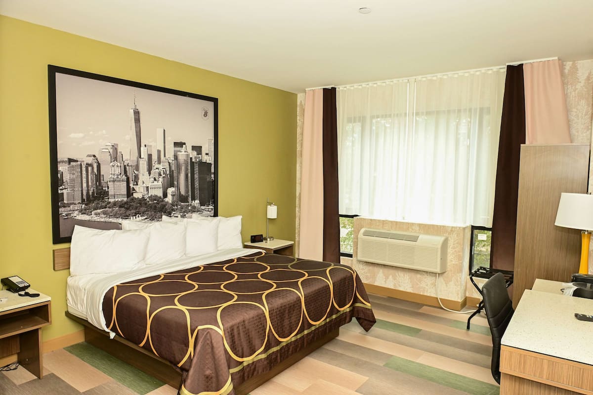 A hotel room with a bed, a desk, a chair, a nightstand, a lamp, a phone, a window with curtains, and a wall-mounted air conditioner.