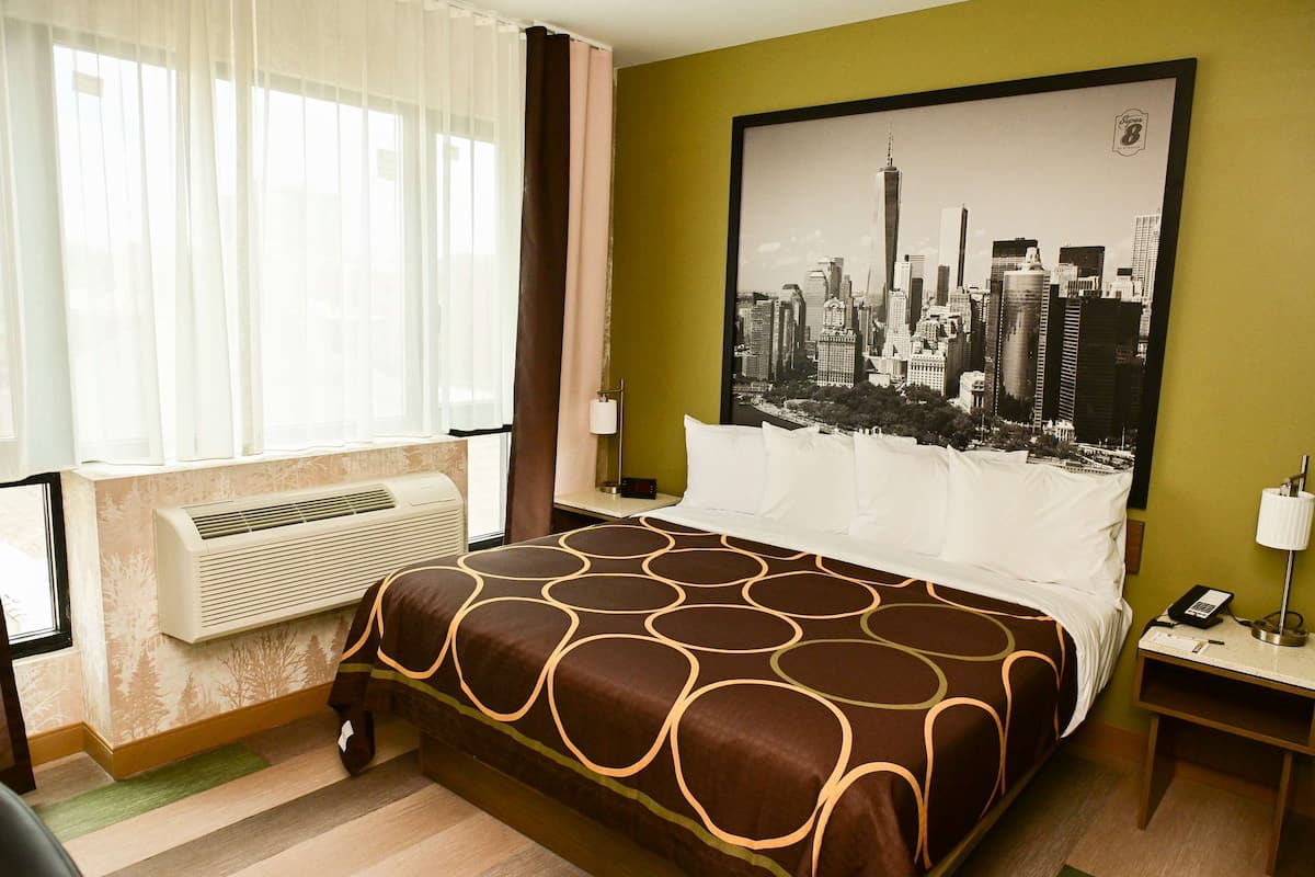 A hotel room with a large bed, a cityscape poster on the wall, a window with sheer curtains, a bedside table with a lamp, and a wall-mounted air conditioner.