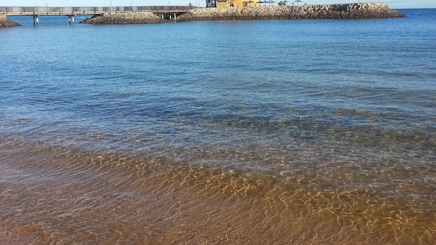 Paisatge costaner amb vista clara a la platja, un moll de fusta i un edifici amb teulada.