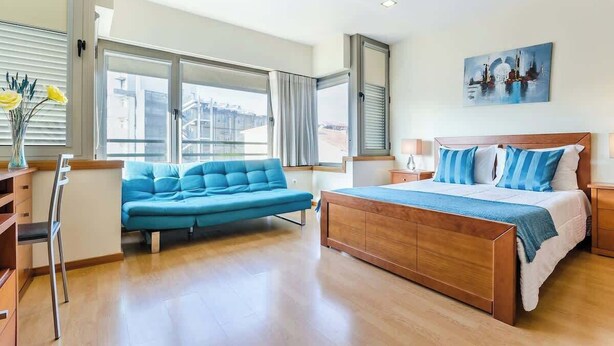 A modern bedroom with a large bed, a blue sofa, and a city view through large windows. - Bright Quiet Flat | Terrace by HostWise (Porto)