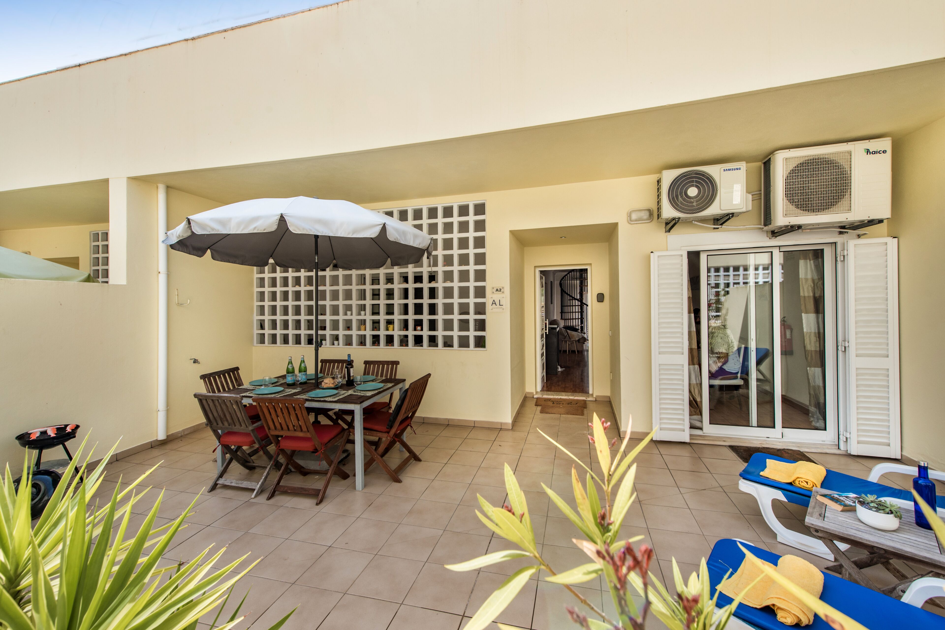 Een terras met een eettafel, stoelen, een parasol, airconditioningsapparaten en een glazen deur die naar een balkon leidt.