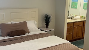 Habitación de hotel con dos camas, una mesita con una planta, y un baño con espejo y artículos de aseo.
