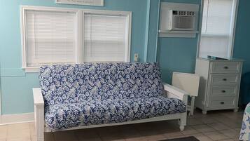 A room with a blue sofa, white dresser, and a window with blinds.
