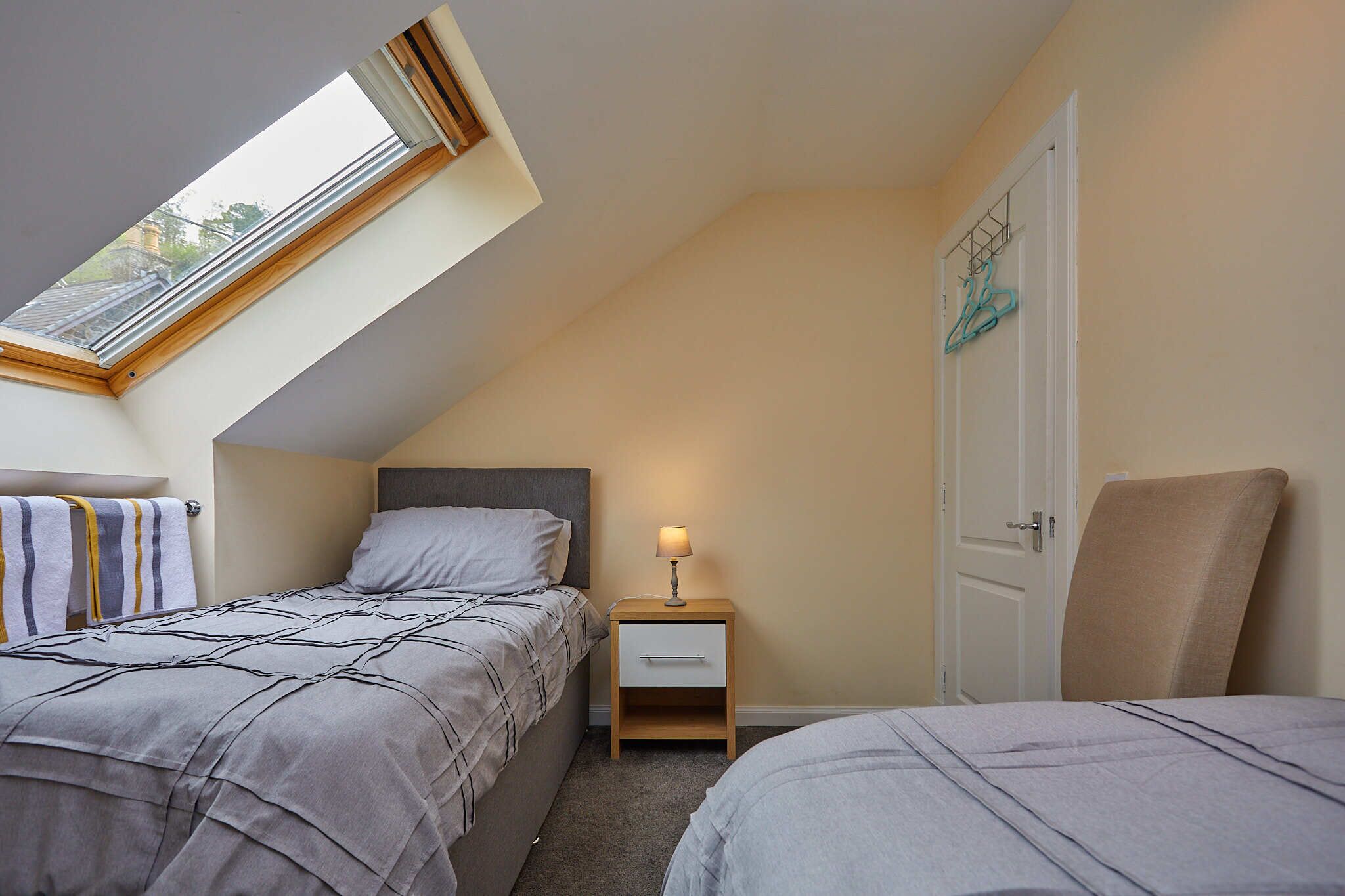 Une chambre avec deux lits, une lucarne, une table de chevet avec une lampe et une porte dotée d’un porte-serviettes.