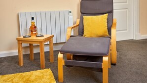 A modern living room with a grey cushioned chair, a small wooden table with a bottle and glasses, and a radiator. - Campbells Loft (Oban)