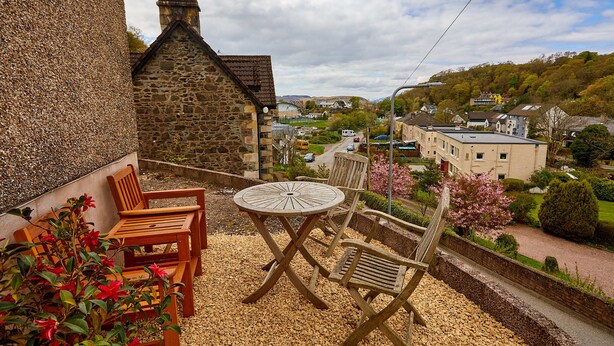 A patio with a round table and wooden chairs, overlooking a village with houses and greenery. - Campbells Loft (Oban)