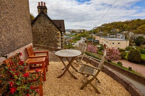 A patio with a round table and wooden chairs, overlooking a village with houses and greenery. - Campbells Loft (Oban)