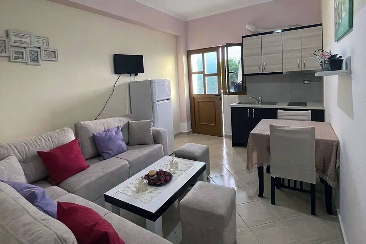 A living room with a sofa, ottoman, and a coffee table. There is a TV mounted on the wall and a kitchen area with cabinets and a sink.