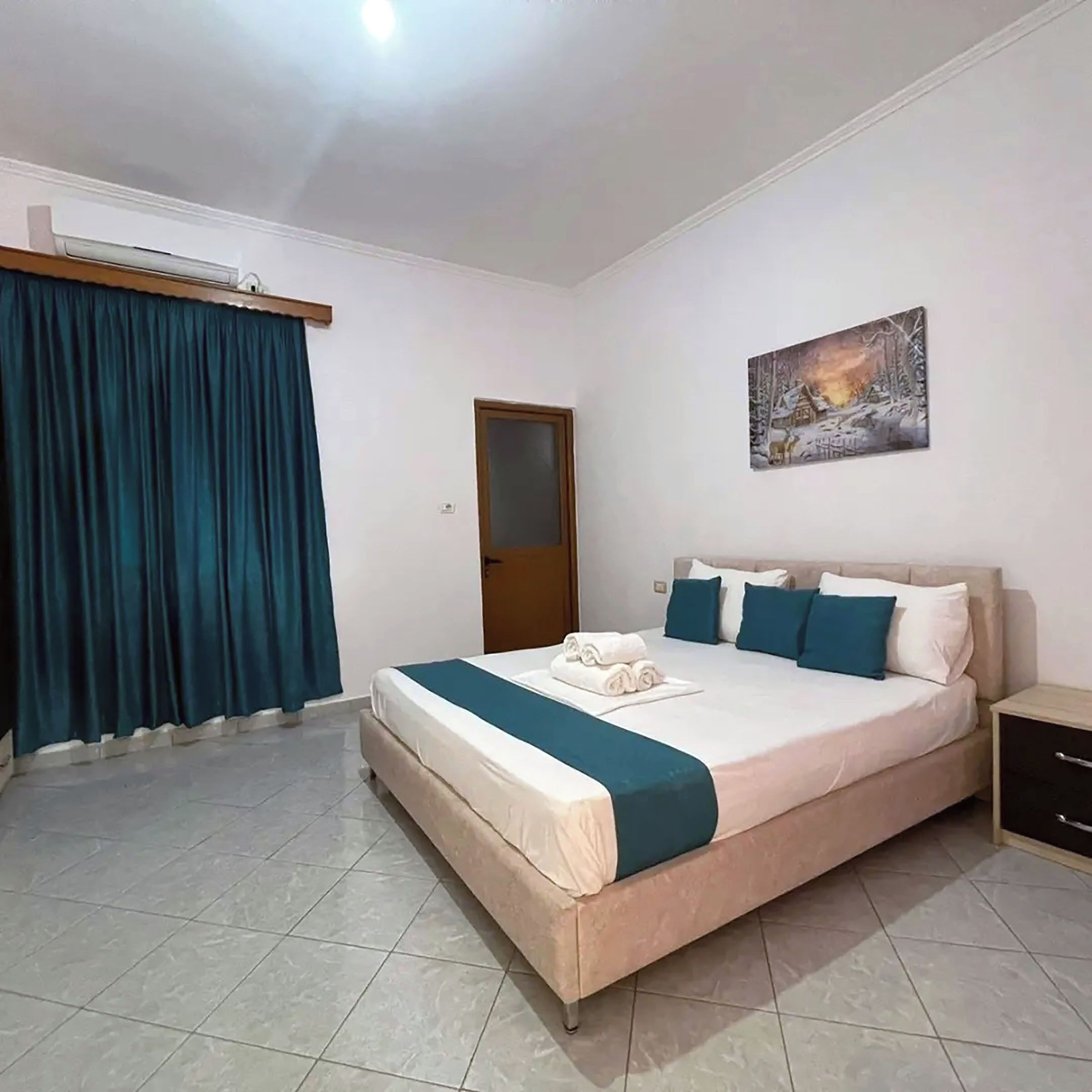 A hotel room with a bed, a nightstand, a painting, and a window with curtains.