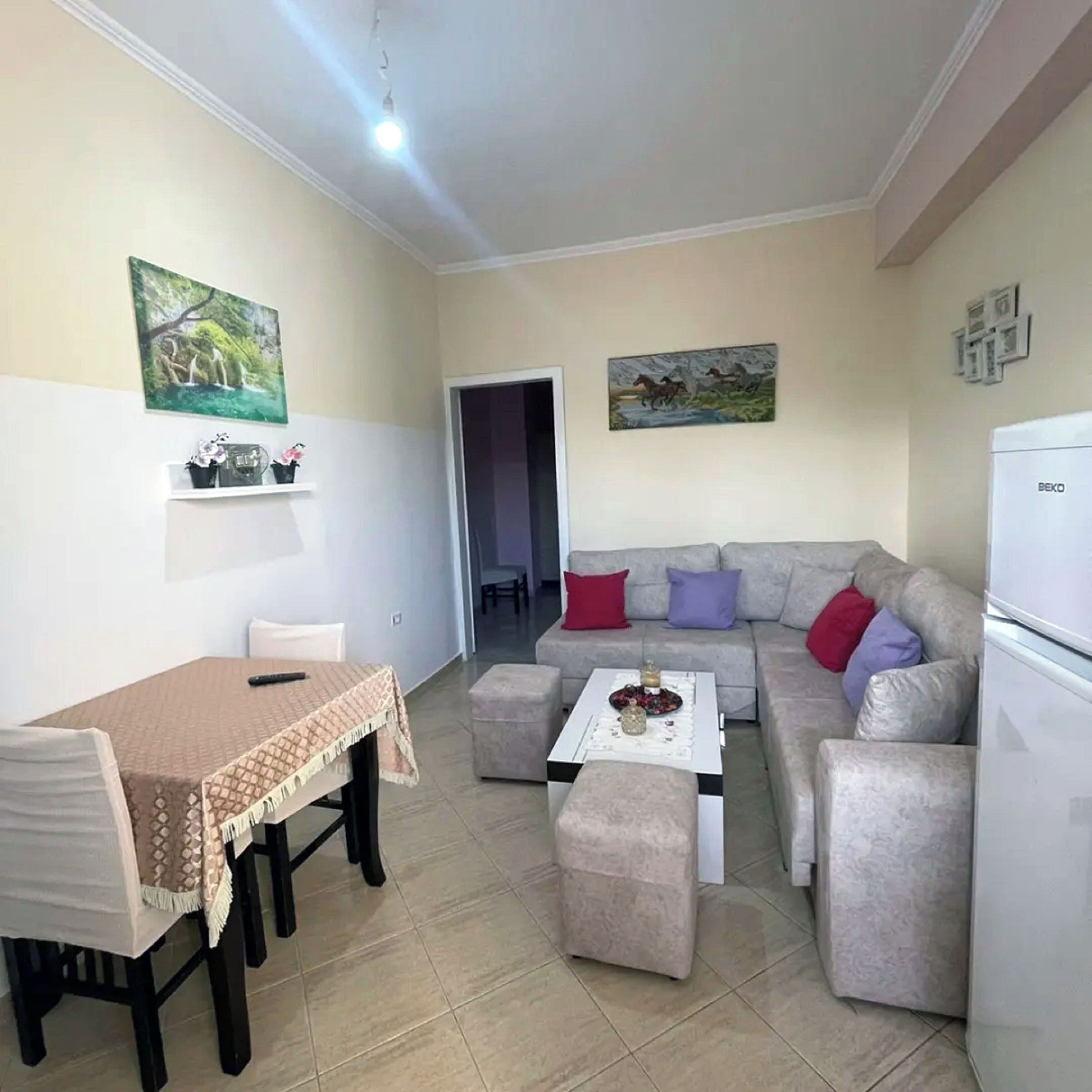 Una sala de estar con una mesa de comedor, sofá y un refrigerador. Hay dos cuadros en las paredes y un estante pequeño con objetos decorativos.
